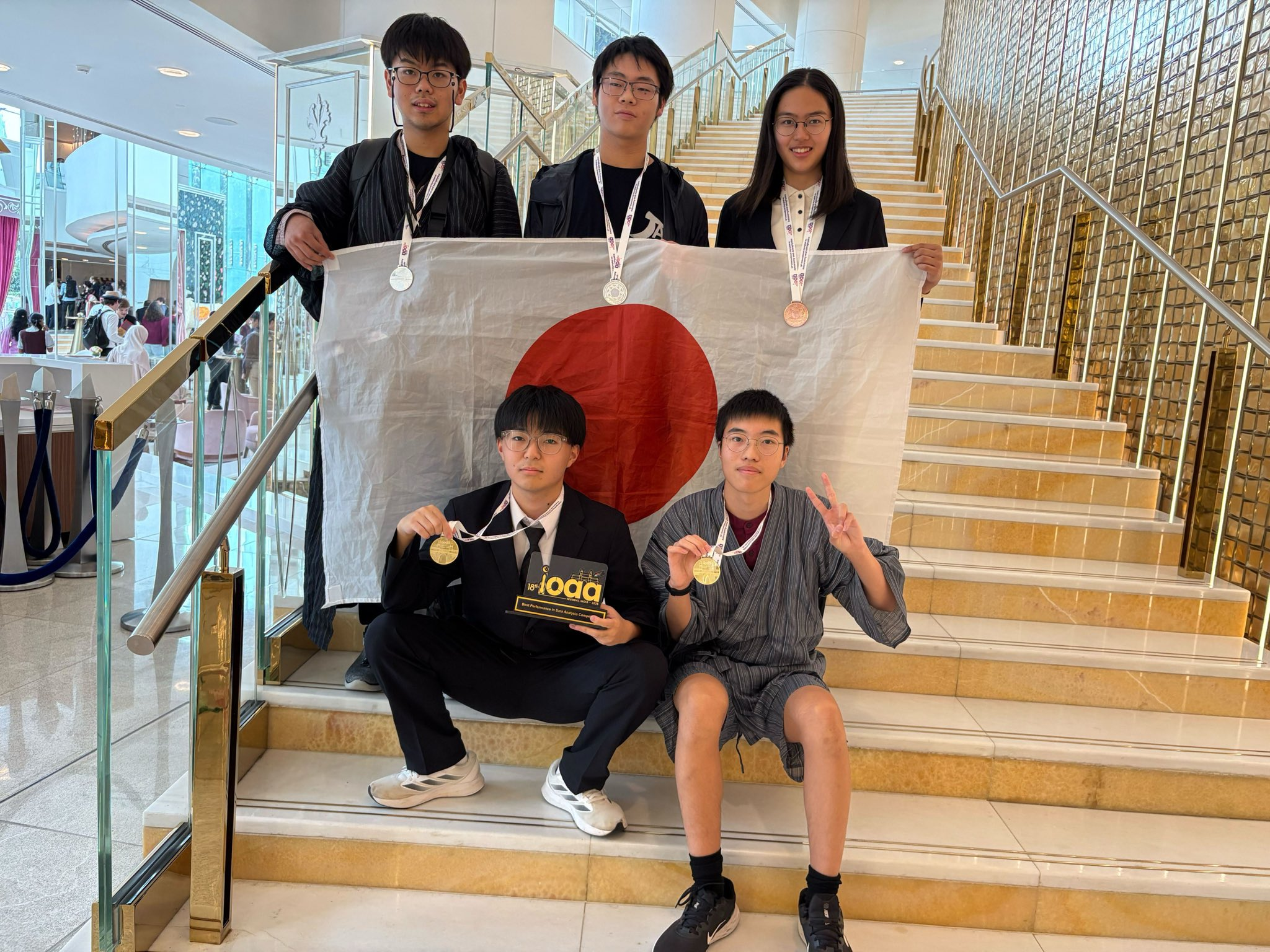 IOAA2025日本代表団の集合写真。階段に立つ生徒5人が写っており、全員がメダルをかけている。後列の3人は日本国旗を持っている。